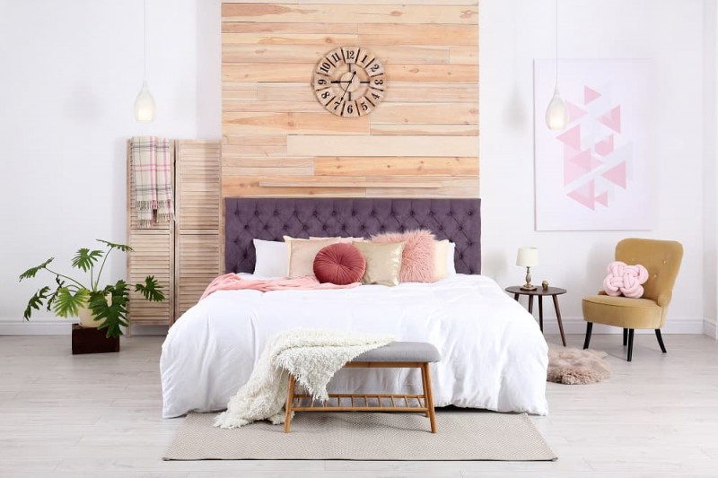 biała sypialnia z drewnianą ścianą za ł&oacute;żkiem i fioletowym zagł&oacute;wkiem